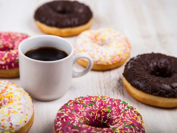 A white cup of coffee surrounded by colorful sprinkled donuts on a wooden surface.