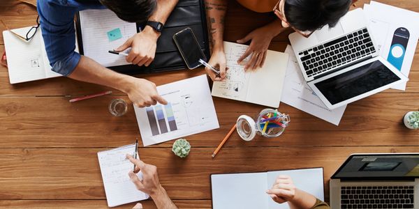 Four people collaborating over charts, notes, and laptops at a wooden table.