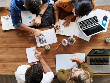 Four people collaborating over charts, notes, and laptops at a wooden table.