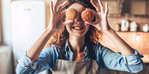 Woman playfully holding two muffins over her eyes, smiling in a cozy kitchen.