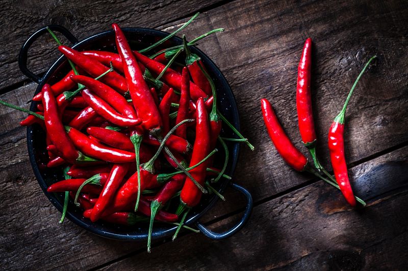 Red chili peppers shot from above on rustic wooden table. A chili peppers heap is in a small pan and three chili peppers are out placed directly on the table. Predominant colors are red and brown. Low key DSRL studio photo taken with Canon EOS 5D Mk II and Canon EF 100mm f/2.8L Macro IS USM