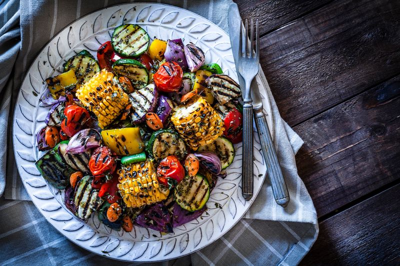Close-up view of a grilled vegetables plate shot from above on rustic wooden kitchen table. The plate is on a gray textile napkin at the left of an horizontal frame and a fork and knife are at the right of the plate. DSRL studio photo taken with Canon EOS 5D Mk II and Canon EF 100mm f/2.8L Macro IS USM