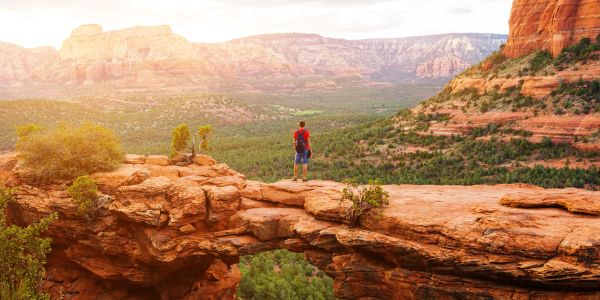 A person standing on a natural rock arch overlooking a valley.