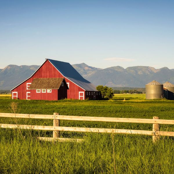 A red barn and silos on a green farm field with mountains in the background.
