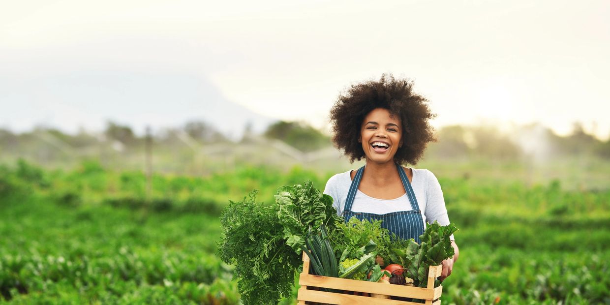 Happy woman holding a crate of fresh vegetables outdoors.