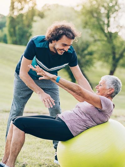 Mature woman exercising on ball outdoors with trainer helping.