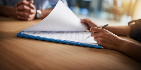 2 people sitting at a table. Just hands on table. One is looking through paperwork with a pen 