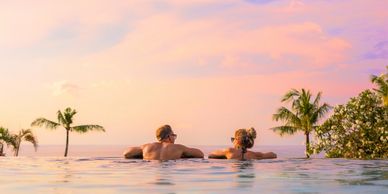 Couple relaxing in an infinity pool at sunset with palm trees around.