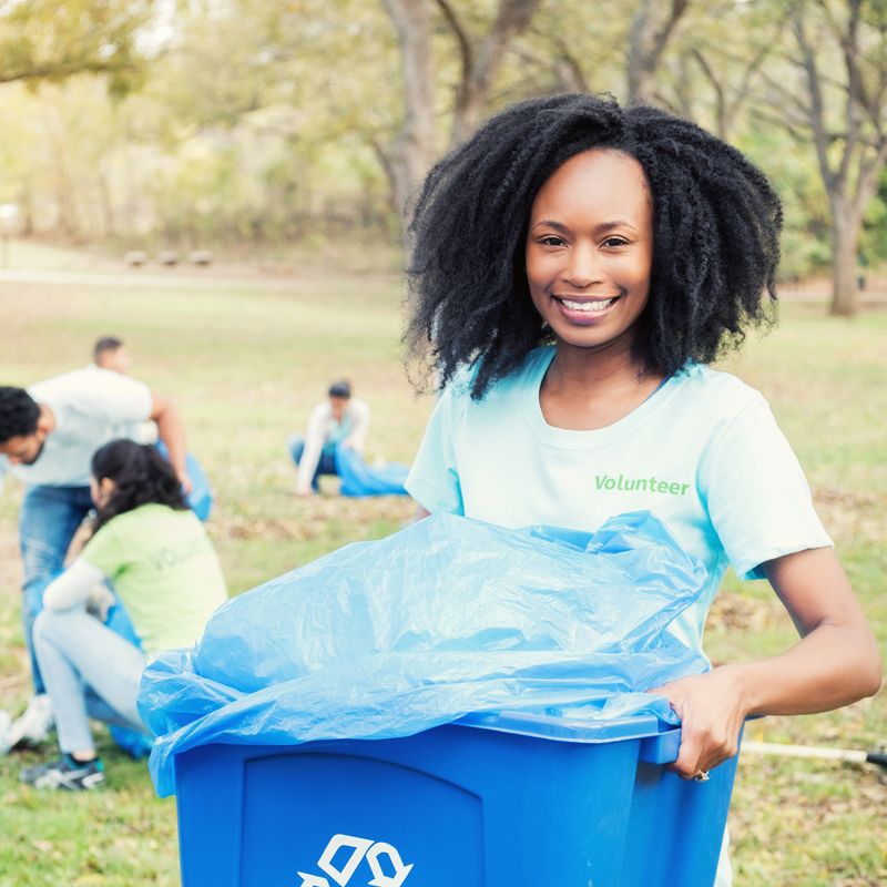 A beautiful African American woman smiles for the camera as she stands outdoors holding a recycle bin.  She wears a volunteer t-shirt as she helps with a community cleanup project.