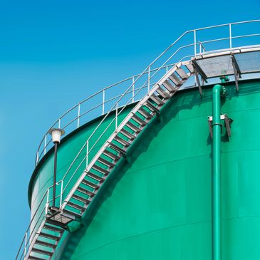 Curved metal staircase on a large green industrial tank under a clear blue sky.