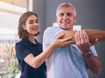 A therapist assists an older man with arm stretches in a bright room.