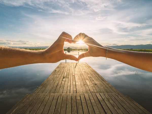 Hands forming a heart shape over a serene lakeside dock at sunset.