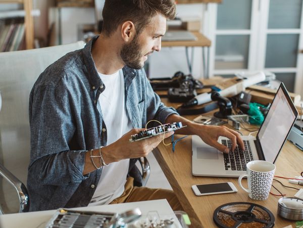 Man working on electronic circuit board with laptop in workspace.
