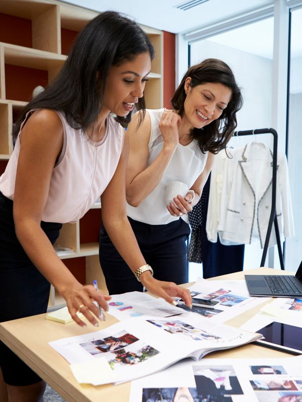 Two women collaborating on a fashion project around a table with sketches and photos.