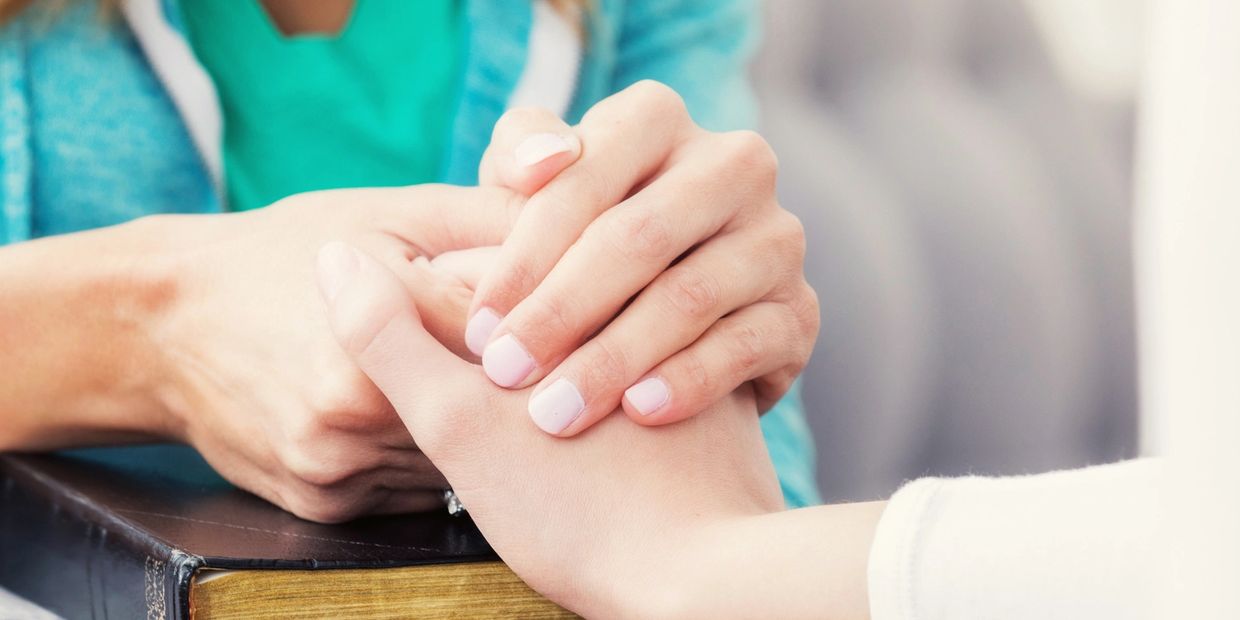 Two women praying and holding hands over a Bible. 
