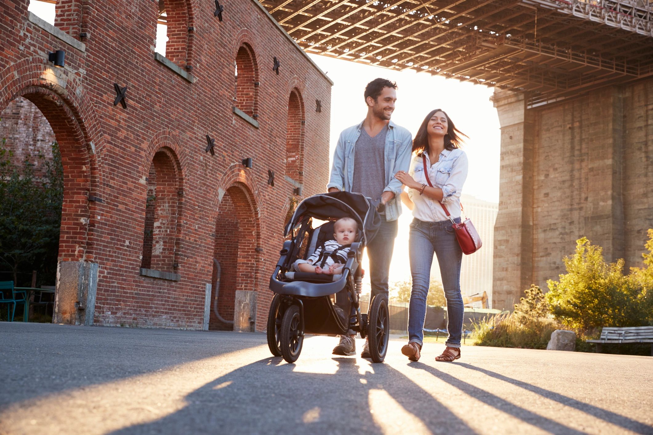 Happy couple walking with baby stroller under a bridge on a sunny day.