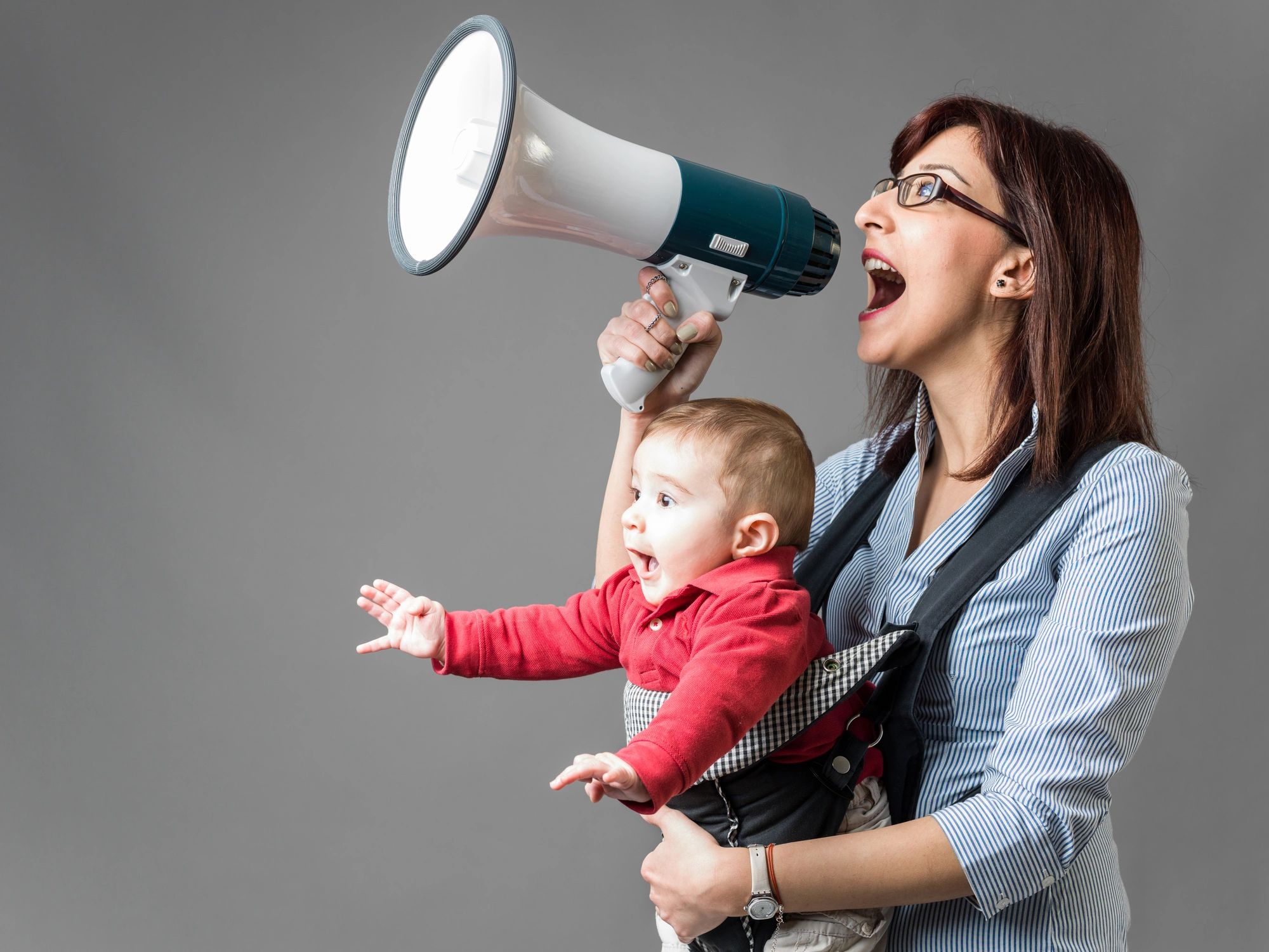 Young mother carrying her baby in baby carrier and shouting through megaphone in front of gray background. Baby is wearing a red t-shirt and he is shouting and excited too. Shot in studio with a medium format camera.