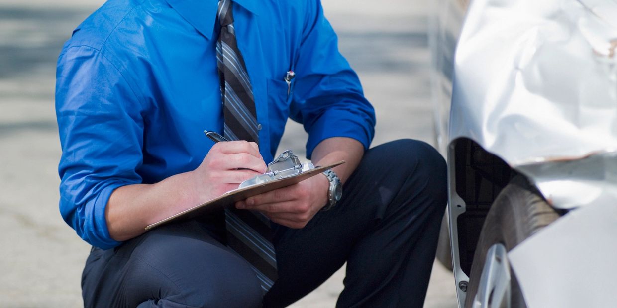 Man in blue shirt inspecting and documenting car damage with a clipboard.