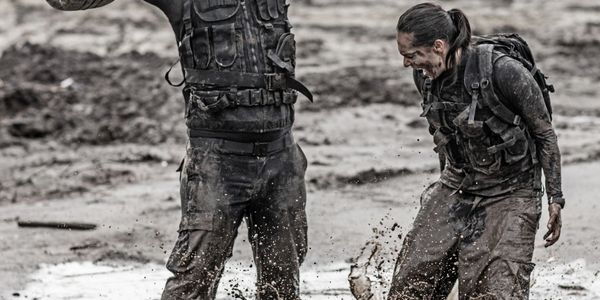 Two people in tactical gear training in muddy terrain, one lifting a wooden bar with tires.