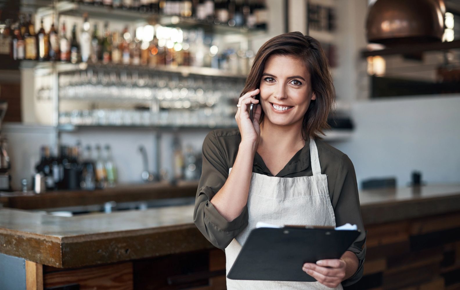 Smiling woman in apron talking on phone, holding clipboard in a bar.