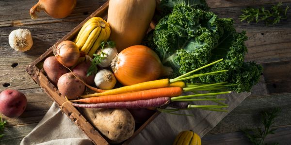 A wooden crate filled with fresh vegetables including carrots, onions, and kale.