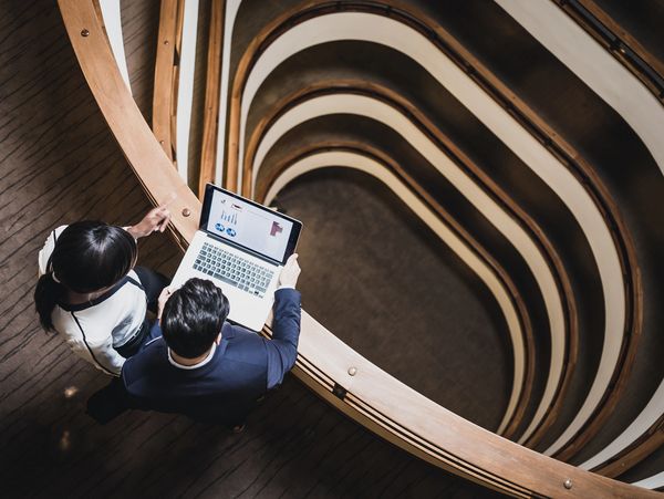 Two professionals reviewing charts on a laptop by a spiral staircase.