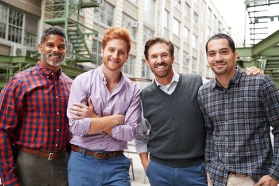 Four men standing together outdoors in casual attire, smiling.
