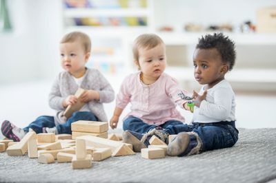 Three toddlers playing with wooden blocks on a carpeted floor indoors.