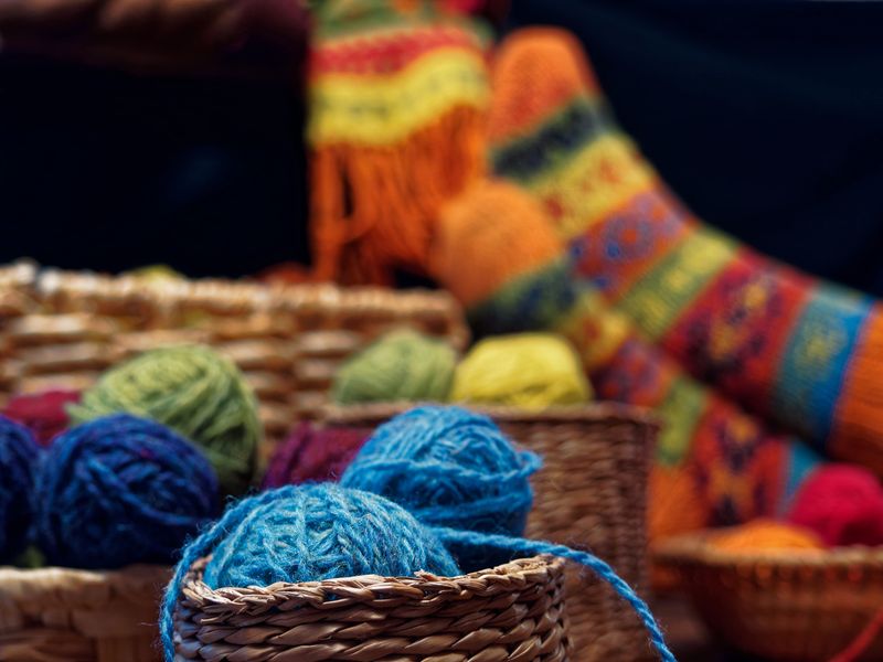 Feet of person wearing winter hand-knitted socks sitting at the table covered with multi-colored balls of wool and knitwear