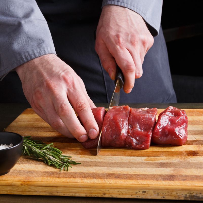 Male chef slicing filet mignon at open restaurant kitchen. Fresh meat, garlic and rosemary for marinade on wooden board. Modern cuisine backgroung with copy space