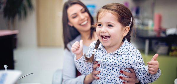 Happy little girl giving thumbs up with smiling woman behind.