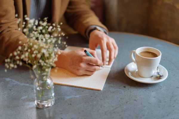 Person writing on paper at a cafe table with coffee and flowers.