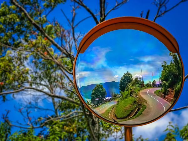 Curved mountain road reflected in a roadside convex mirror under a blue sky.