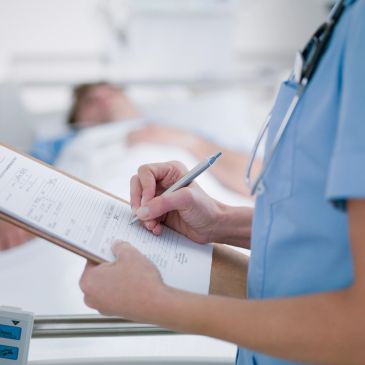 Nurse writing on clipboard near patient bed.