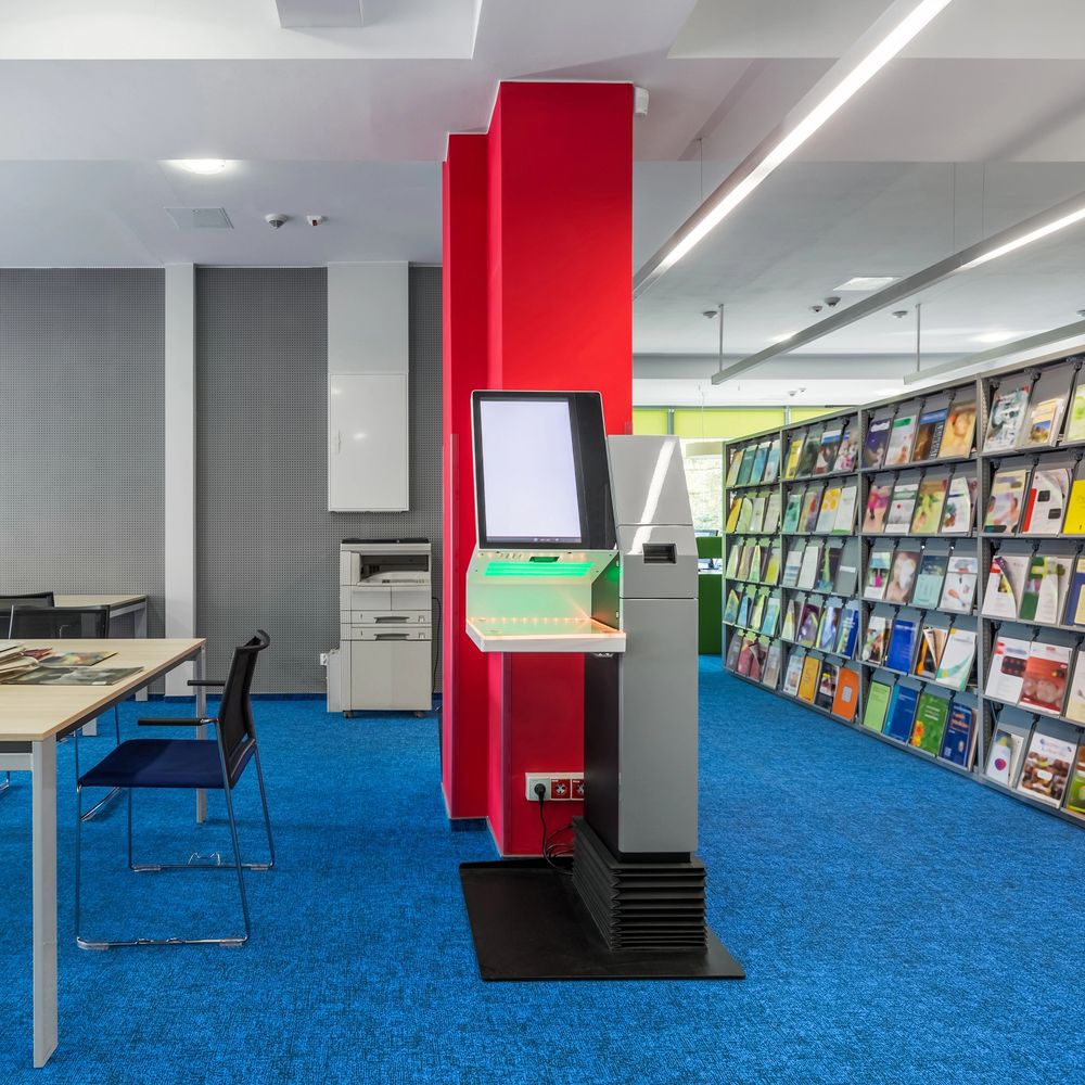 Modern library interior with a self-service kiosk, blue carpet, and shelves of magazines.