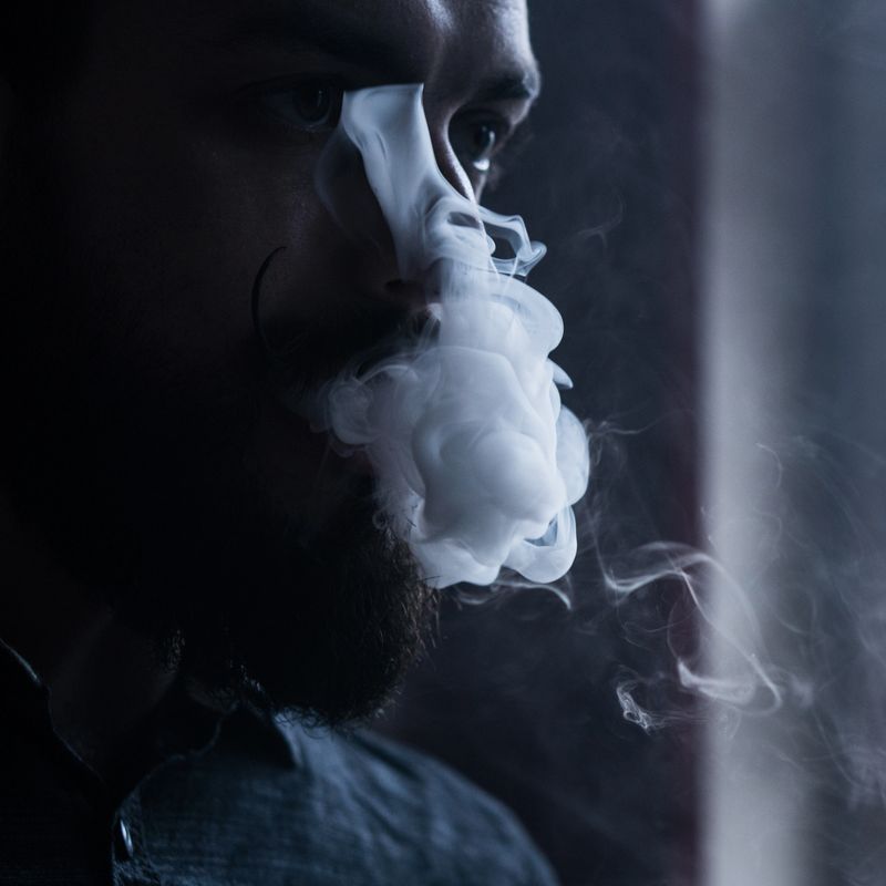 Young Man with Beard and Mustages Vaping an Electronic Cigarette. Vaper Hipster Smoke Vaporizer and Exhals Smoke Flow.