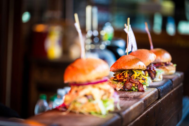 Close up image of a selection of freshly flame grilled burgers in a row on a wooden counter at Borough Market, one of the oldest and most famous food markets in the world. Each of the burgers has its own label, on which is written the contents of the burger. The burgers are sandwiched between glazed buns, and presented on beds of fresh green lettuce and stuffed with melted cheese and red onion. Horizontal colour image with copy space.