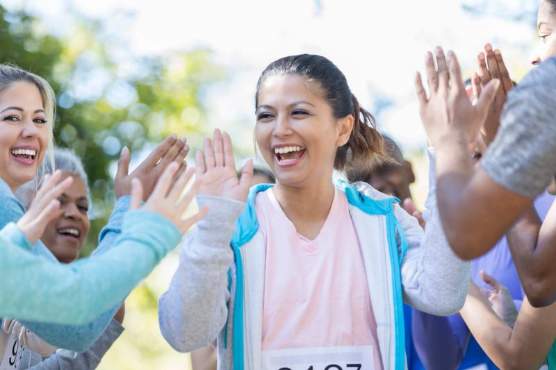 A laughing mid adult woman uses both hands to give high fives to a crowd of people after finishing a charity race.