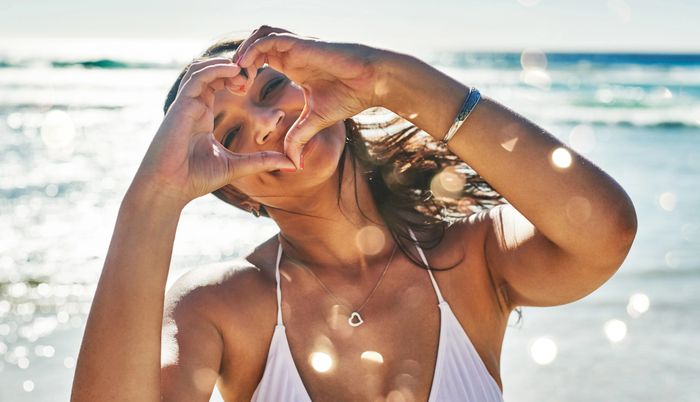 A woman forming a heart shape with her hands on a sunny beach.