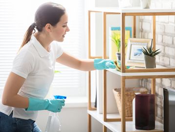 Woman cleaning a shelf with gloves and spray bottle.