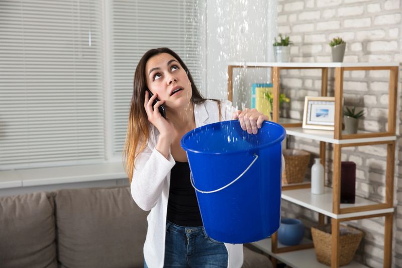 Worried Woman Calling Plumber While Collecting Water Droplets Leaking From Ceiling At Home
