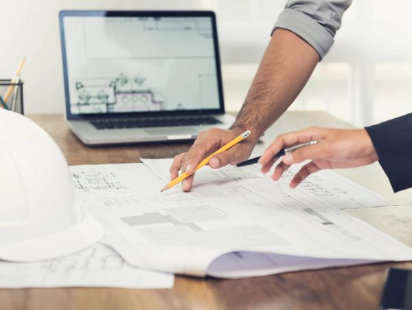 Two people discussing architectural blueprints at a desk with a laptop and hard hat.