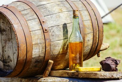 Wooden barrel with a corked bottle of amber liquid and two wrapped items on a wooden surface outdoors.