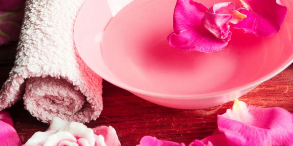 Pink bowl with water and rose petals beside a rolled towel on wooden surface.
