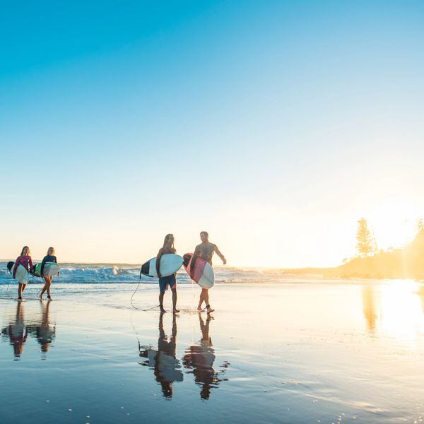 Four surfers walk along a reflective beach at sunset with their surfboards.