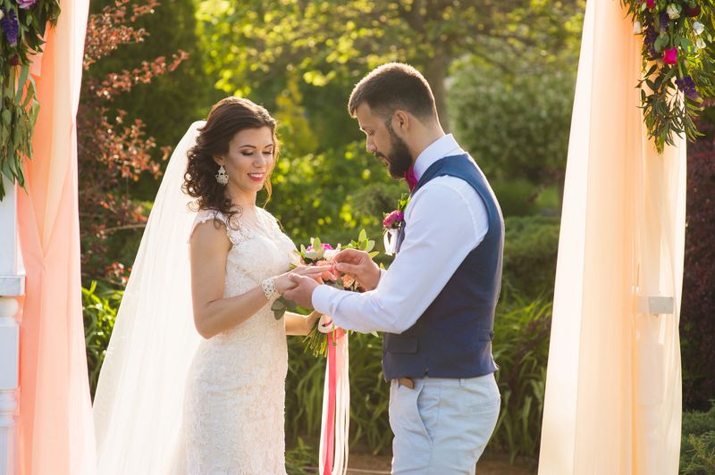 Groom wear ring in the finger of his bride unter the arch with peach color textile