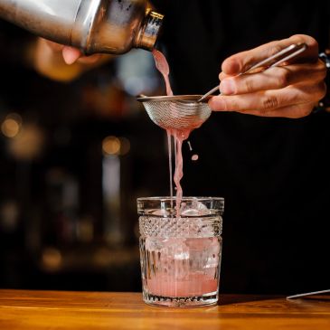 Pouring a pink cocktail through a strainer into a glass with ice.