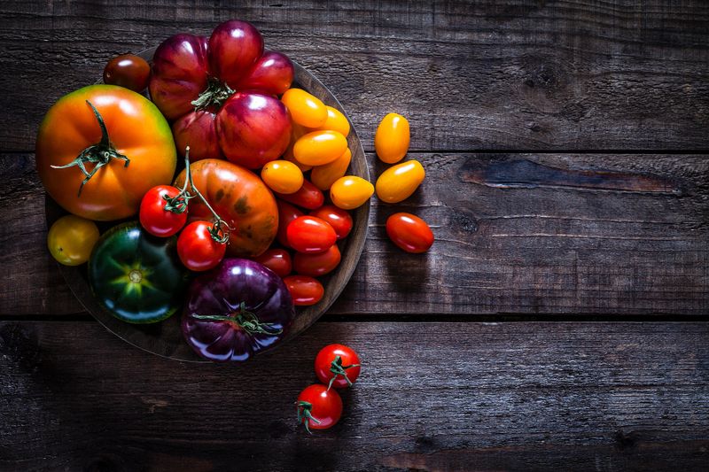 Variety of multi colored ripe tomatoes shot from above on rustic wooden table. The tomatoes are grouped on a wooden cutting board at the left of an horizontal frame leaving useful copy space for text and /or logo at the right. Low key DSRL studio photo taken with Canon EOS 5D Mk II and Canon EF 100mm f/2.8L Macro IS USM