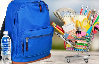 Blue backpack and mini shopping cart filled with school supplies on a table.