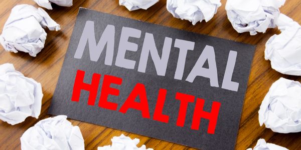 Card with 'MENTAL HEALTH' surrounded by crumpled paper balls on wooden surface.